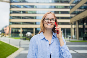 Vertical shot of happy young blonde woman makes telephone call in roaming wears casual clothes had glad expression walks against blurred city building enjoys smartphone talking. High quality photo