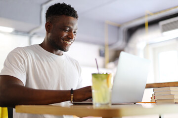 Happy African American man who is typing on his laptop in a bright cafe. He is a young and casual student, businessman or entrepreneur who uses technology and the internet to study.