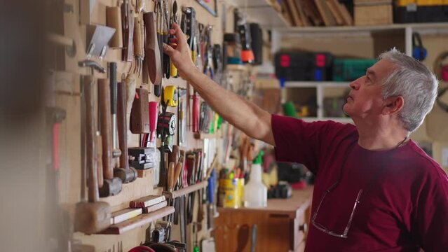 Older man working at workshop, grabbing tools from wall and engaged with craftsmanship work hobby. Senior elderly worker