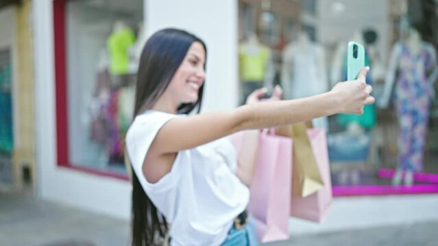 Young beautiful hispanic woman make selfie by smartphone holding shopping bags at clothing store