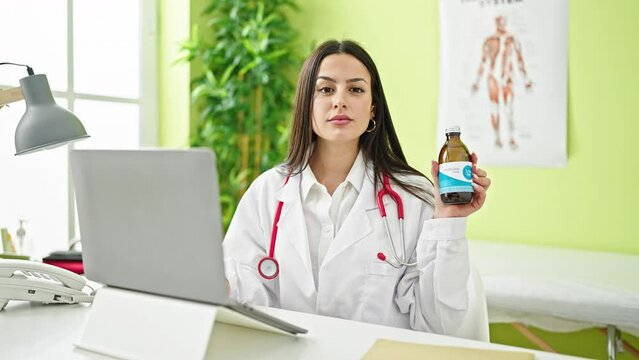 Young beautiful hispanic woman doctor using laptop holding medication bottle at clinic