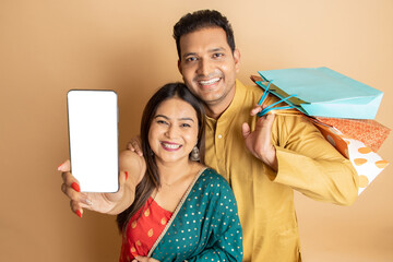 Happy young indian couple wearing traditional cloths holding shopping bags and and smart phone with blank display screen isolated on beige background. Diwali celebration and festive offer and sale.