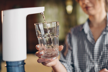 A woman pours water from a cooler into a glass in the kitchen at home. Close up