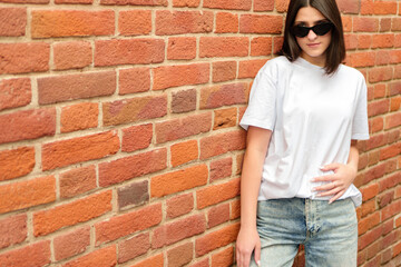 Obraz premium Summer portrait of a brunette girl in sunglasses against a brick wall with a place for text. City life concept.