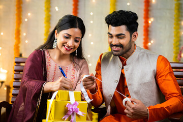 Happy indian married couple busy preparing gift box by writing names during festival ceremony - concept of love, caring or sharing present and traditional culture