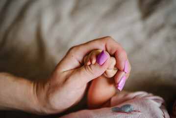 Baby hand. Young mother holding child's hands. Cute little baby lying on bed at home. Mom hold son fingers. Closeup. Tiny hand of newborn baby girl or boy wrapped in hands. Cute sleeping daughter.
