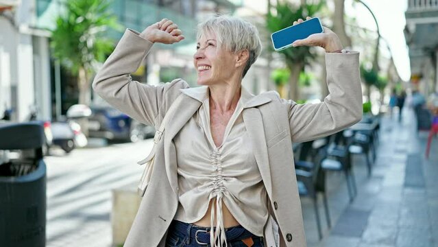 Middle age blonde woman smiling confident using smartphone with winner expression at coffee shop terrace