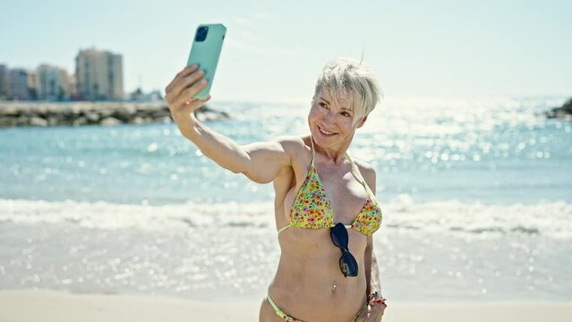Middle age blonde woman tourist wearing bikini make selfie by smartphone at the beach