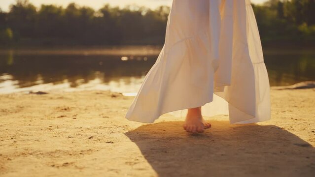 women's legs feet close-up walk along beach river banks, stepping barefoot on yellow sand. Woman enjoys summer nature free life water green tree sun light. Girl walks go in white long old style dress