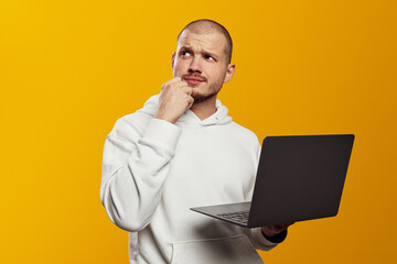 Thoughtful young bearded man holding laptop computer and touching chin with hand and looking away, isolated over yellow background - obrazy, fototapety, plakaty