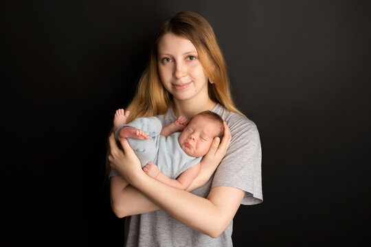 A cute newborn baby boy sleeps in a blue overalls in the first days of life. In the arms of the mother. Warm motherly hugs. On a black background. Professional portrait photography.