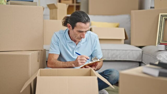 Middle age man checking cardboard box writing on clipboard at new home