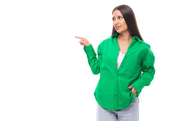 attractive young caucasian brunette lady with makeup dressed in an elegant green shirt points her finger on a white background with copy space