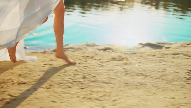 women's legs feet close-up runs walks along beach river banks, stepping barefoot on yellow sand. Body part happy Woman enjoys nature free life water sun light. Girl go in white long silk dress train