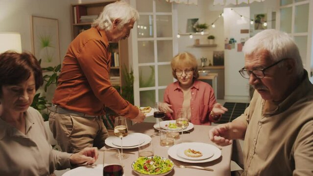 Medium shot of senior couple treating friends with festive dishes sitting together at table at cozy home birthday celebration party