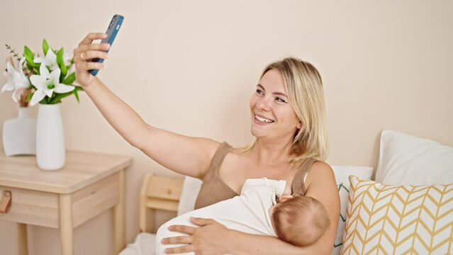 Mother and daughter sitting on bed breastfeeding baby make selfie by smartphone at bedroom