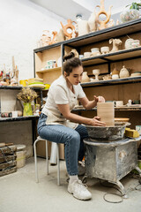 Brunette craftswoman in apron creating clay vase on pottery wheel in ceramic studio