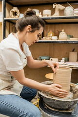Side view of craftswoman in apron working with clay vase on pottery wheel in studio in workshop