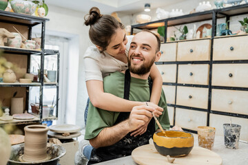 Joyful potter embracing boyfriend coloring clay bowl and working in ceramic studio at background