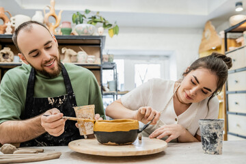 Cheerful craftspeople in aprons coloring clay bowl while working together in ceramic workshop