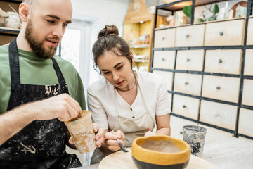 Craftsman in apron coloring clay bowl  with blurred girlfriend together in ceramic workshop