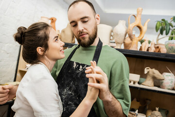 Bearded sculptor in apron holding hand of smiling girlfriend while working in ceramic workshop