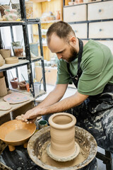 Bearded sculptor in apron washing hands in bowl with water near clay and pottery wheel in workshop