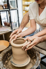 Fototapeta na wymiar Cropped view of potter molding clay with girlfriend on pottery wheel in ceramic workshop