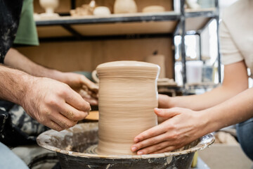 Fototapeta na wymiar Cropped view of romantic artisans shaping clay vase on pottery wheel together in ceramic studio