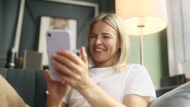 Portrait of charming blond mature woman hold smartphone scrolling watching social media indoors Happy female texting on her phone enjoying leisure time at home