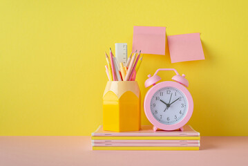 Back to school concept. Photo of school accessories on pink desktop alarm clock stand for pencils over stack of copybooks and sticky note paper attached to yellow wall