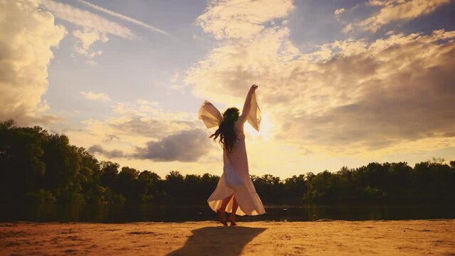 dark silhouette happy woman dancing by river bank water sun light blue sky green forest tree. Fantasy girl raised hand to sky enjoy nature. herbal wreath long hair white dress fly in wind slow motion
