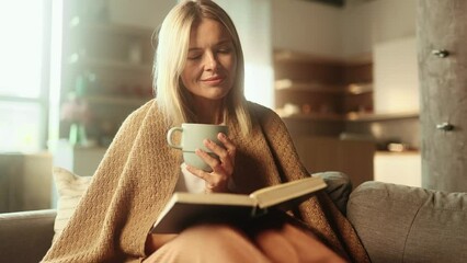 Portrait of charming blond mature woman under the blanket with cup of hot tea or coffee and reading book literature at home Calm relaxed female enjoying weekend leisure time alone indoors