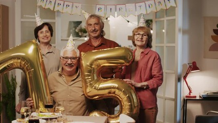 Portrait shot of happy pensioners posing for camera with balloons in shape of number seventy five when celebrating anniversary at home party