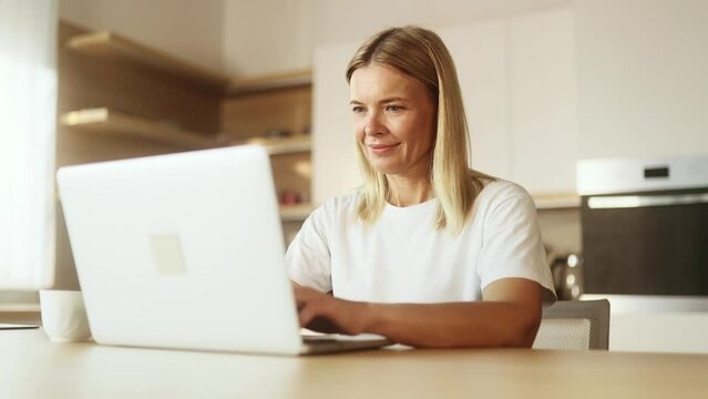 Portrait of blond mature woman working online typing browsing products in internet store reading news checking email or social media profile on laptop computer at light home indoors