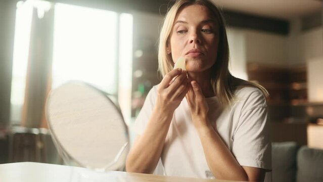 Portrait of pretty blond mature woman looking at mirror reflection and doing self massage with gouache moon stone at home Concept of skincare cosmetology natural beauty cosmetics face building