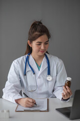 Young beautiful asian woman doctor holding pills bottle writing on document at clinic