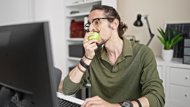 Young hispanic man business worker using computer eating apple at office