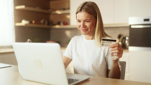 Portrait of charming mature blond woman paying with credit card on laptop at home Happy customer doing payments online shopping in internet store and receiving cash back indoors 