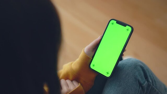 Hand of woman holding mobile smartphone and swipes photos or pictures with blank green screen mock up while sitting on a sofa at home.