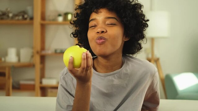 Happy pretty girl biting green apple at home. Beautiful african american young woman eating fresh fruit and smiling. Healthy food vegan vegetarian dieting concept. Healthy snack clean food