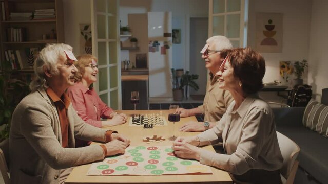 Medium shot of positive pensioners sitting at big table and figuring out who they are in guessing game at home party