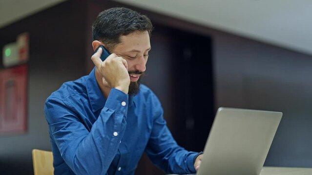 Young hispanic man business worker using laptop talking on smartphone at the office