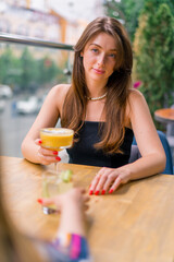 a young smiling girl with a cocktail in her hands sits and chats with a friend at a table on the summer terrace overlooking the city in a bar club