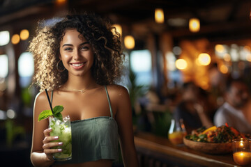 An attractive young woman holds a refreshing drink with a mint leaf on top, mojito, she is in a...