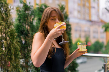 Portrait of a young smiling girl with cocktails in her hands and drinking one of them on the summer terrace in a club bar 