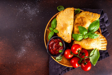 Homemade patty pies, baked pastries with meat or vegetables. Italian calzone closed pizza, Mexican empanada, Karaim Ukrainian pies. With homemade tomato sauce, basil greens and spices on dark table