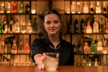 portrait of a smiling girl bartender who puts a cocktail with ice on the bar and serves it to the...