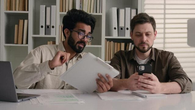 Two businessmen multiracial men co-workers in office Arabian Indian businessman show papers business project on laptop to colleague Caucasian man ignoring addicted mobile phone inefficient teamwork