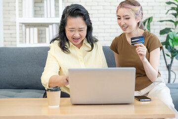 Smiling asian mother and cute daughter are happy while making online shopping successfully by laptop in living room at home enjoying leisure together. Online shopping concept